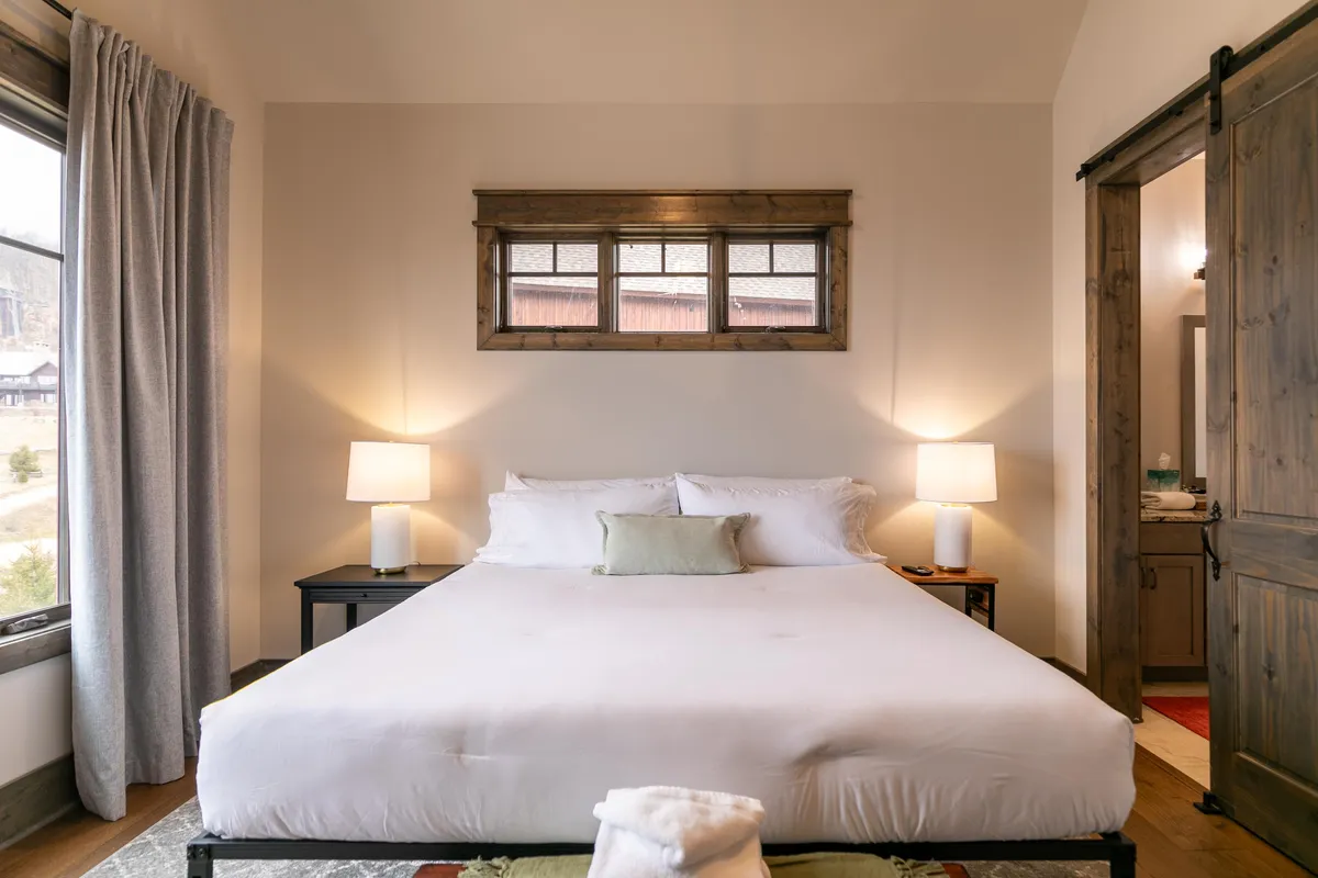 A serene king-sized bed with plush bedding, flanked by bedside lamps, sits beneath a large window with wooden trim. The natural light softly illuminates the room, creating a tranquil atmosphere ideal for relaxation or reading.