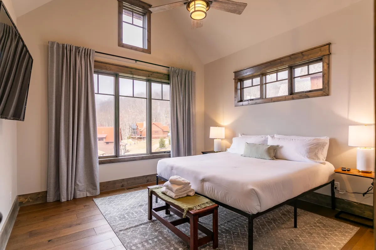 A cozy bedroom featuring a neatly made bed, soft rug, nightstands, and a generous window that lets in abundant natural light. The scene evokes peaceful mornings and quiet escapes with a view of the outdoors.