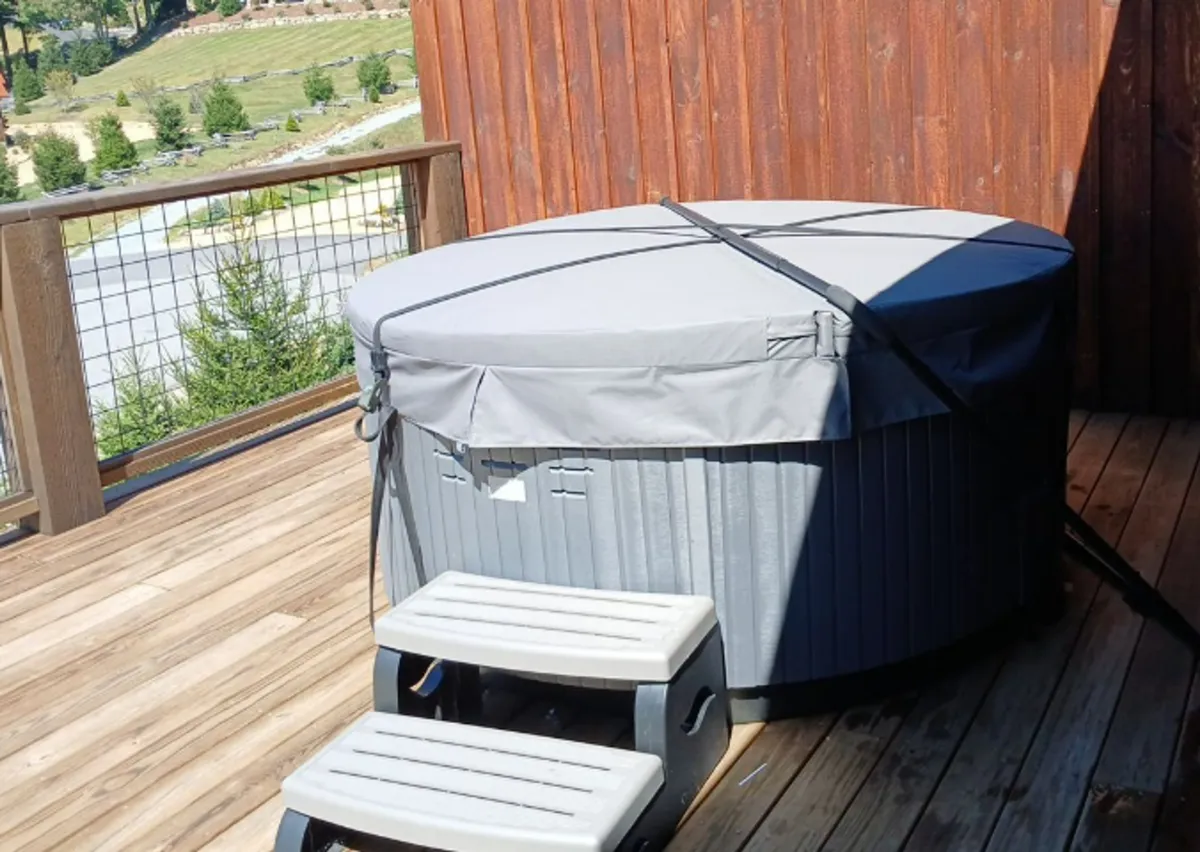 A tranquil outdoor wooden deck featuring a bubbling hot tub nestled among lush greenery. The hot tub is the focal point, with surrounding natural scenery offering a serene, private atmosphere perfect for relaxation. The image captures the seamless blend of comfort and nature, with the deck space inviting for both soaking and lounging.