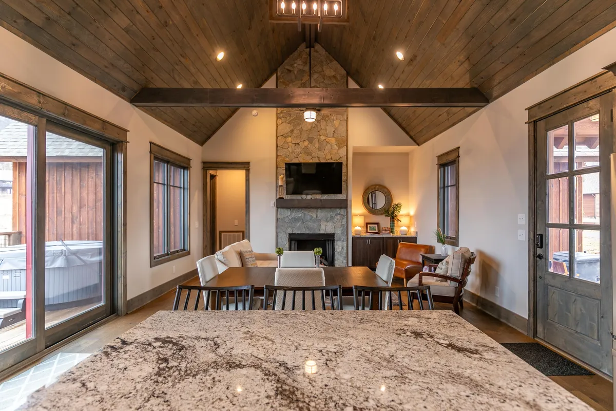 This image displays an inviting living and dining space with a granite countertop, a stone fireplace, exposed wooden beams, and large windows that flood the room with natural light. The area is arranged for family gatherings, casual dining, or simply relaxing by the fire, offering a warm, communal ambiance.
