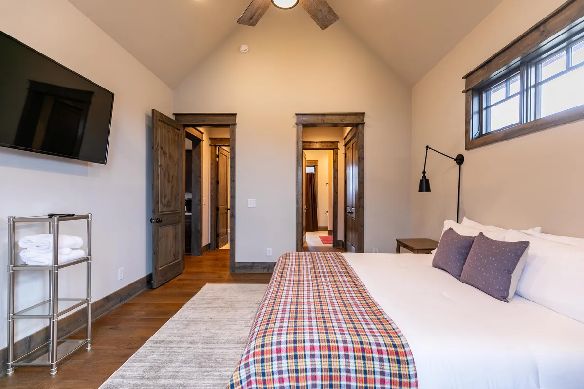 A comfortable bedroom with a bed dressed in a plaid throw, a wall-mounted TV, and doors leading to other rooms. A shelf with towels and soft lighting complete the cozy, contemporary mountain look.