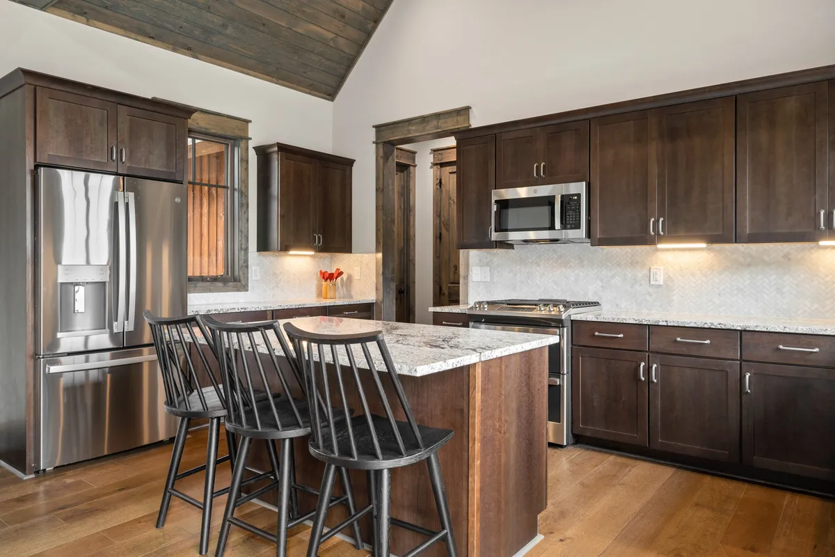 A functional, stylish kitchen featuring a granite countertop, black bar stools, stainless steel appliances, and wooden cabinets. The setup invites both cooking and socializing, with morning sunlight streaming in.
