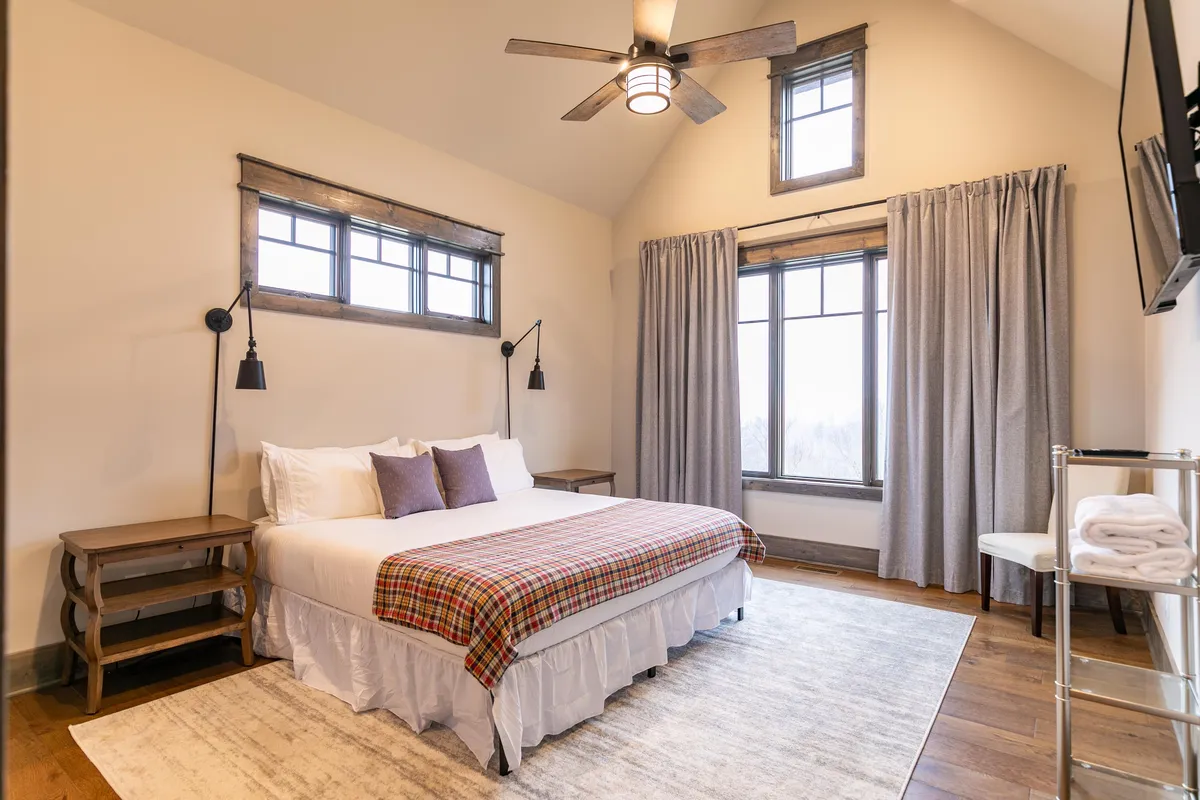 A bedroom scene featuring a bed draped in a plaid blanket, large windows letting in generous natural light, and cozy nightstands with lamps on either side. The space evokes calm, restfulness, and an inviting retreat for guests.